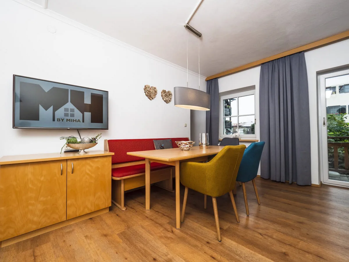 Wohnzimmer mit TV, Tisch und bunten Stühlen, Holzfußboden und Fenster mit Vorhängen.